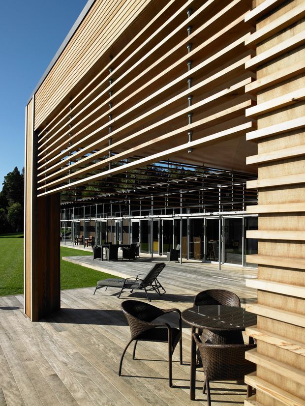 Exterior architectural view of a contemporary multi-level building with extensive timber horizontal slat screening and a deep covered outdoor terrace. Full-height glazing opens onto a timber deck furnished with dark woven outdoor chairs, loungers, and a round table. The open slatted facade creates strong linear shadows and passive shading, with landscaped lawn visible around the building.