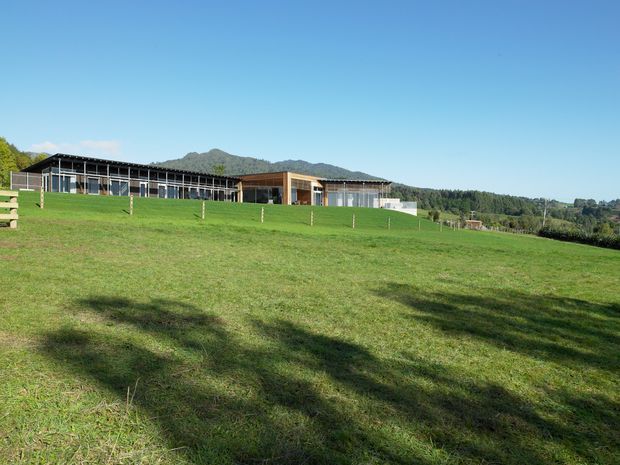 A wide exterior view of a low, contemporary rural property set behind a broad green lawn and fenced paddock. The building has long horizontal glazed wings, dark slender roof and framing elements, and a warm timber-clad central section. Rolling hills and tree-covered terrain form the background beneath a clear blue sky.