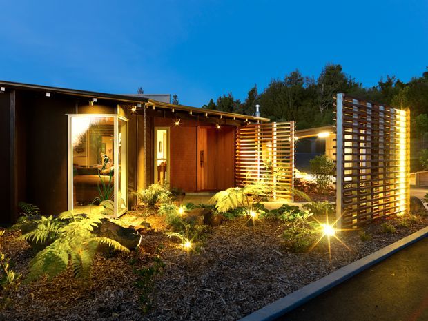 A low, single-storey contemporary building photographed at dusk, with dark cladding, warm timber entry doors, illuminated glazing, and horizontal timber privacy screens. The landscaped frontage includes ferns, rocks, mulch, and integrated ground lighting, creating a warm garden approach.