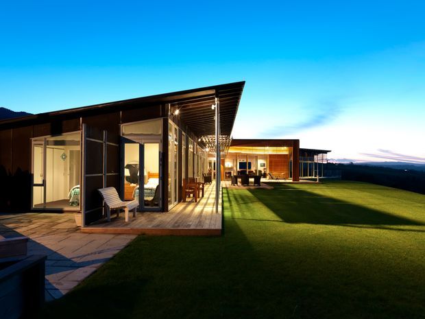A modern single-storey residential building photographed at dusk, with long low wings, extensive floor-to-ceiling glazing, dark timber or metal-clad exterior walls, and broad projecting roof eaves. A covered timber deck and paved terrace connect the interior living spaces to a large manicured lawn. Warm interior lighting contrasts with the cool blue evening sky and distant horizon.
