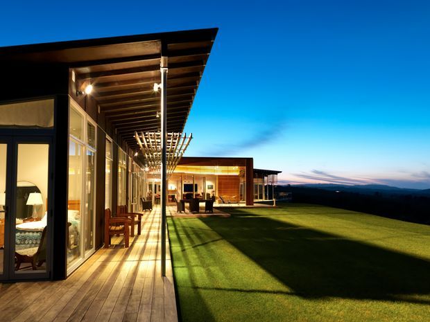 A contemporary single-storey residence photographed at dusk, with a long timber deck and glazed living spaces beneath a deep projecting roof and exposed timber rafters. A pergola extends along the facade, while warm interior lighting reveals furnished rooms through the glazing. The house opens onto a broad manicured lawn with distant dark hills visible beyond.