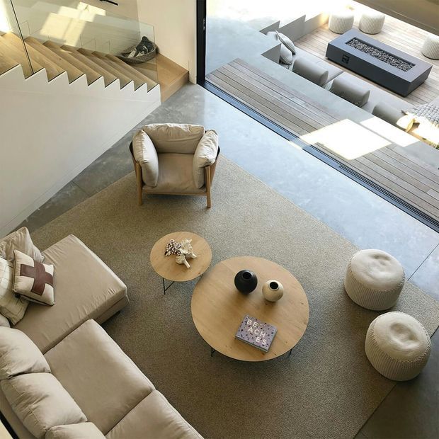 A bright, contemporary living room viewed from above, with a large neutral sectional sofa, a beige lounge chair, two round light-wood coffee tables, textured ottomans, and a muted area rug. A floating timber staircase with a glass balustrade runs along the left wall, while full-height glazing opens onto a timber outdoor deck with a built-in rectangular fire table and outdoor seating.