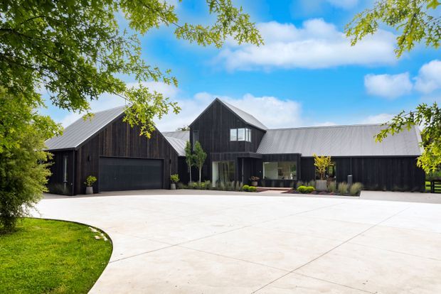 A low, expansive modern residence with dark vertical timber cladding and multiple pitched roof volumes, set behind a broad pale concrete driveway. The central entrance is recessed beneath a projecting volume, with large glazed openings and restrained landscaping. A dark garage door is visible on the left, while mature leafy trees frame the property.