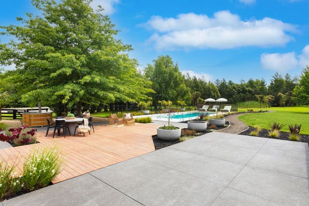 A spacious landscaped outdoor entertaining area with a timber pool deck, outdoor dining furniture, a built-in circular spa or hot tub, and a swimming pool beyond. Large concrete-paved areas lead through manicured lawns, garden beds, mature trees, and poolside umbrellas under a bright blue sky.