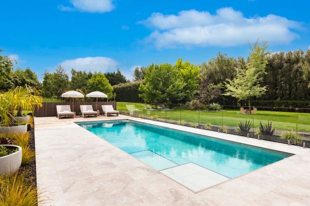 A bright outdoor swimming pool area with a long rectangular turquoise pool, pale stone paving, glass safety fencing, sun loungers and umbrellas, surrounded by manicured lawn, hedges and mature garden planting under a blue sky.