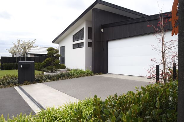 A contemporary residential house exterior viewed from the landscaped front approach, featuring a crisp white rendered facade, dark vertical cladding and trim, a broad white sectional garage door, and a grey concrete driveway. Low-maintenance planting, clipped shrubs, a black mailbox, and a linear drainage channel frame the entry.