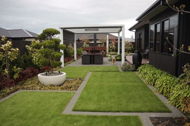 A landscaped residential courtyard viewed from a lawn path, featuring geometric sections of manicured grass bordered by dark paving strips, layered garden beds, a sculpted potted tree, and a contemporary covered outdoor entertaining pavilion with dining furniture. A dark-clad house with large windows lines the right side, while fencing and neighbouring rooftops are visible beyond.