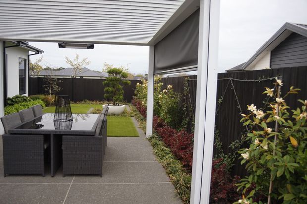 A covered outdoor dining terrace with a dark wicker dining table and chairs, concrete paving, and a retractable louvred roof. The compact landscaped courtyard includes lawn, raised planting beds, climbing and flowering plants, and dark privacy fencing. Adjacent suburban homes are visible beyond the boundary.