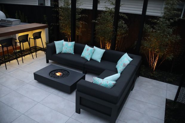 A stylish outdoor entertaining area with a black sectional lounge arranged around a square gas fire-pit table on large light-grey tiled paving. A compact bar with stools sits at the rear, backed by dark screening and illuminated bamboo planting. The space has a refined, intimate evening atmosphere.