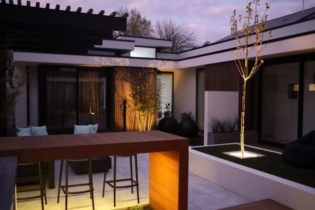 A contemporary courtyard-style outdoor entertaining area at dusk, featuring a long timber bar with stools, integrated warm lighting, dark-framed glazing, rendered walls, raised garden beds, and a small illuminated feature tree. The low, layered rooflines and pergola create a sheltered modern courtyard atmosphere.