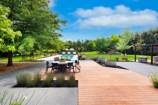 A spacious landscaped outdoor entertaining area with a large timber deck, dining table and chairs, and a swimming pool beyond. Concrete paving, raised garden beds, ornamental grasses, mature trees, sculptural planters, and a covered outdoor seating structure create a polished contemporary garden setting.