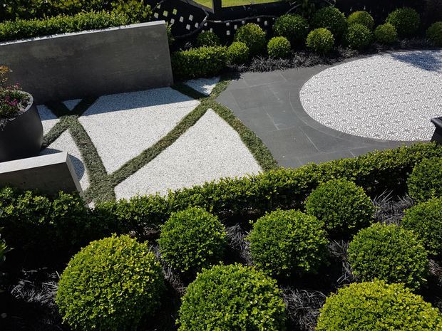 A contemporary landscaped courtyard or garden viewed from above, featuring geometric white gravel or pebble paving divided by narrow planted strips, dark grey rectangular paving, a large circular patterned pebble area, clipped spherical shrubs, low hedges, ornamental grasses, and dark rendered raised planters.