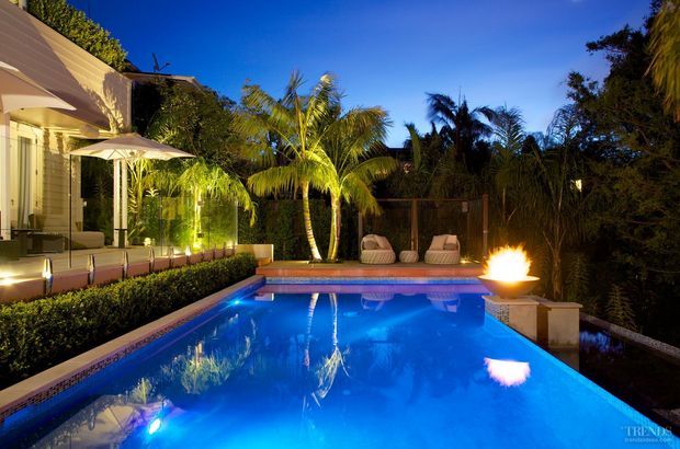 Evening exterior view of a landscaped residential pool area, featuring a bright blue swimming pool, tropical palms, dense planting, illuminated lounge seating, a poolside fire feature, and a covered outdoor entertaining area beside the house.