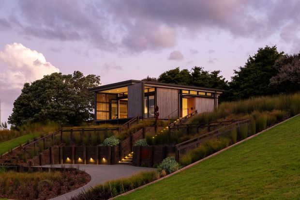 A low-profile contemporary house set into a landscaped sloping site at dusk. The building features extensive floor-to-ceiling glazing, dark vertical cladding, warm interior lighting, and a flat roofline. Terraced garden beds, timber retaining elements, pathway lighting, and native-style planting surround the house.