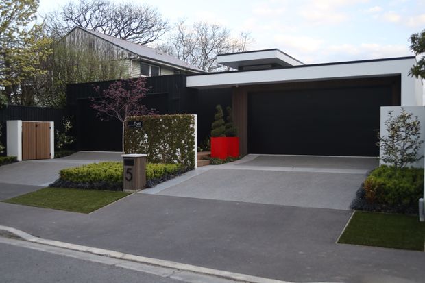 Street-facing view of a contemporary residential property with a dark rendered garage facade, flat-roofed volumes, timber and black screening, concrete driveway, landscaped hedges, and a small pedestrian gate. The house sits in a leafy suburban setting.