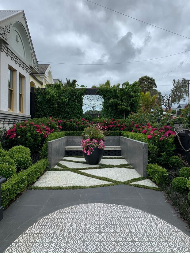 Formal landscaped courtyard garden viewed from a dark paved entry, featuring a patterned circular tile inlay, clipped box hedges, abundant pink flowering shrubs, white gravel paths with grass joints, and a curved concrete bench. A white ornate residential facade with decorative classical detailing is visible at left beneath an overcast sky.