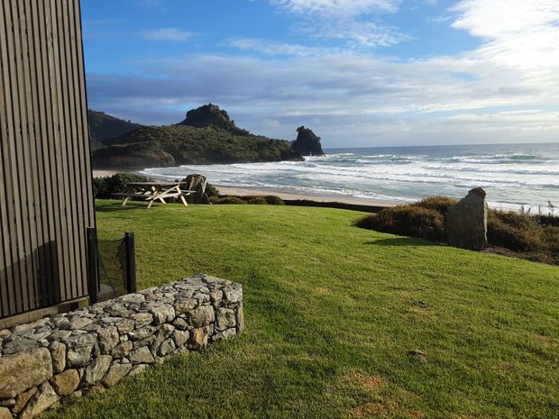 A coastal outdoor view from beside a dark vertically clad building, overlooking a neatly mown lawn, low dry-stone retaining wall, picnic table, sandy shoreline, surf, and rugged rocky headland beneath a partly cloudy sky.