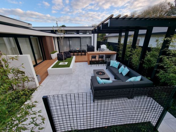 A contemporary courtyard entertaining area viewed from above, featuring pale tiled paving, timber decking, a dark pergola with exposed beams, an outdoor dining table, and a large charcoal outdoor lounge with turquoise cushions. The courtyard is enclosed by modern white and dark-framed glazed buildings, raised planters, and a black wire fence.