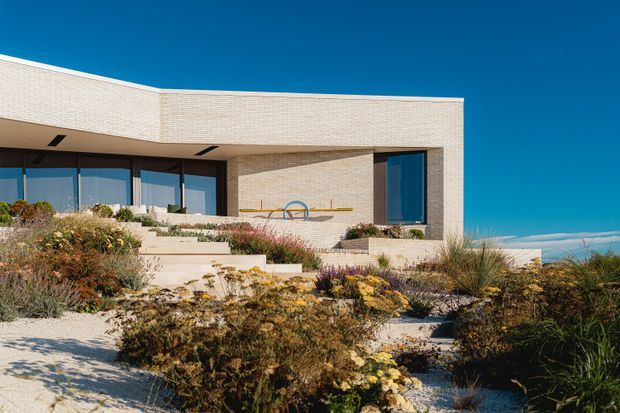 Exterior view of a contemporary, low-profile home with pale textured masonry walls, expansive dark-framed glazing, and a flat roof. The building is set within a landscaped coastal or dune-like garden with sandy ground and naturalistic planting; a blue ocean-like horizon is visible beyond.