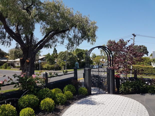 A landscaped residential front entrance viewed from the property side, featuring a decorative black metal arched gate, low masonry boundary wall, patterned paved forecourt, clipped shrubs, flowering roses, and mature street trees beyond. The setting appears suburban and sunny.