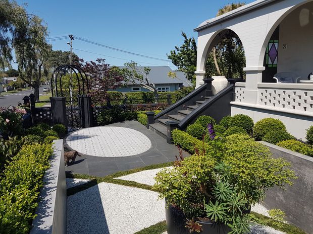 A landscaped front garden and entry courtyard with geometric light paving, dark charcoal paths, clipped hedges, ornamental planting, and a black arched garden gate. The raised house entry features a white rendered facade, arched openings, a decorative breeze-block balustrade, dark steps, and a small stained-glass panel. A suburban street and neighbouring houses are visible beyond the garden.