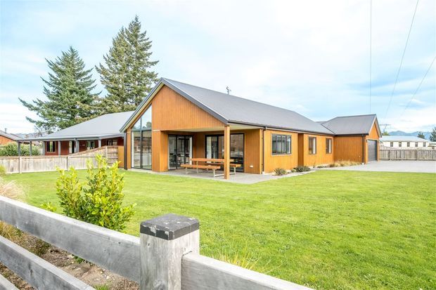 Single-storey contemporary timber-clad house with a prominent pitched gable roof, large glazed entry or living area, covered timber porch, and expansive manicured lawn. The building uses warm vertical timber cladding with dark grey roofing and trim, set in a suburban or semi-rural landscape.