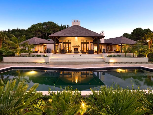 A symmetrical single-storey residence viewed from across a large landscaped swimming pool at dusk. The home features a broad hipped roof with dark shingles, warm cream rendered walls, extensive black-framed glazed doors and windows, a raised terrace with wide steps, and matching side wings. Tropical planting and integrated landscape lighting surround the pool and house.