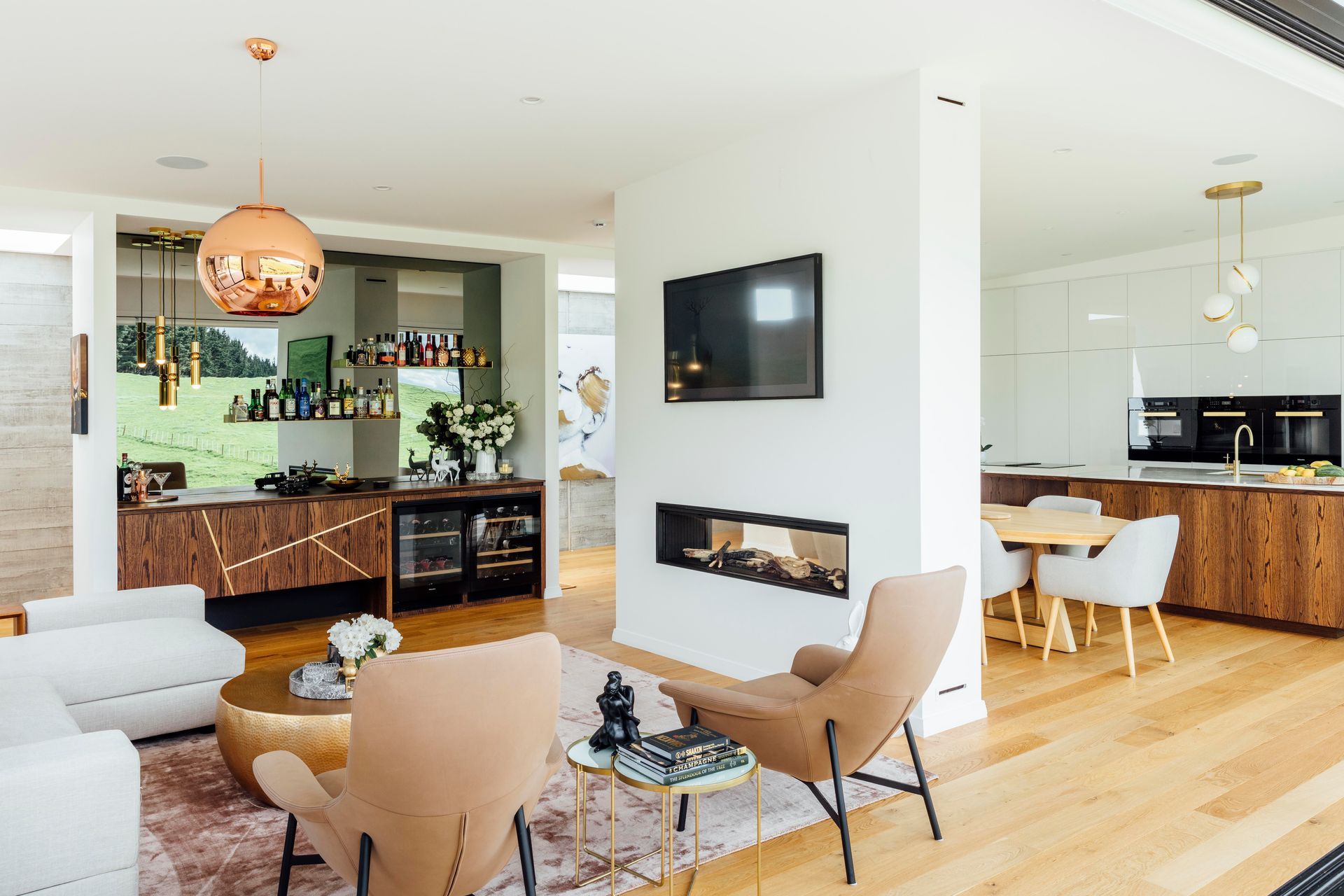 A double-sided fireplace separates the kitchen from the living room and bar where Lee Broom pendants hang above the cocktail cabinetry. A Tom Dixon copper pendant lends metallic magic, and a smokey mirror reflects the view.