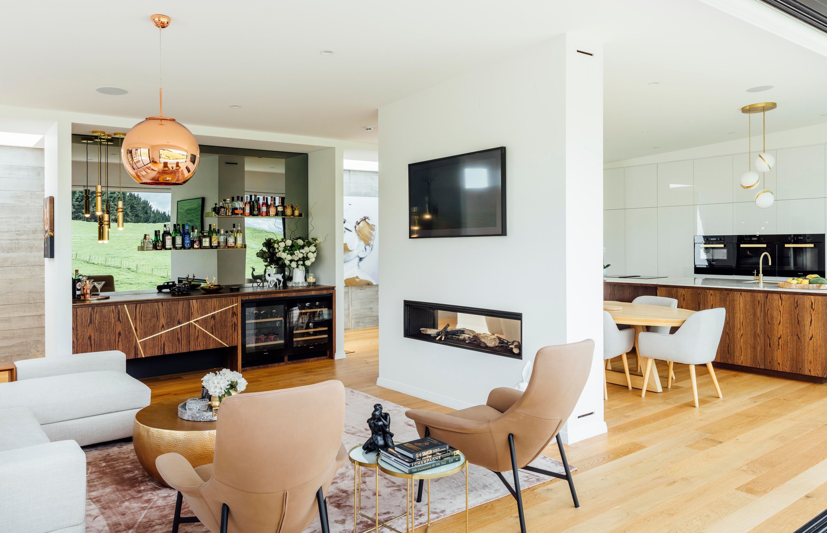 A double-sided fireplace separates the kitchen from the living room and bar where Lee Broom pendants hang above the cocktail cabinetry. A Tom Dixon copper pendant lends metallic magic, and a smokey mirror reflects the view.