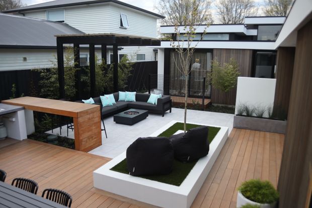 A contemporary courtyard terrace viewed from an elevated interior or deck position, featuring extensive timber decking, a built-in outdoor dining bench and table, black-framed pergola, sectional seating around a fire pit, raised white planter beds with ornamental grasses and a central artificial grass feature. Surrounding architecture uses horizontal weatherboard cladding, dark metal roofing and timber-lined walls with large windows and covered connections.