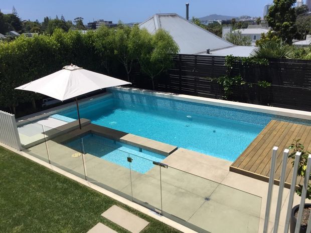 A bright residential backyard swimming pool area with a large rectangular pool, integrated shallow spa or wading section, glass safety fencing, pale concrete paving, timber decking, a white cantilever-style umbrella, and landscaped screening. Low-rise suburban buildings and distant hills are visible beyond the black privacy fence.