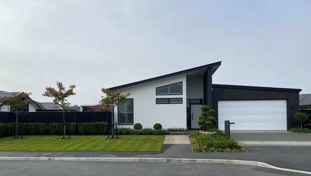 Contemporary single-storey suburban house with a crisp white rendered facade, dark charcoal pitched roof and garage volume, horizontal windows, landscaped front lawn, clipped shrubs, and a simple concrete path leading to the recessed entry. The composition emphasizes the home's street-facing exterior and clean geometric form.