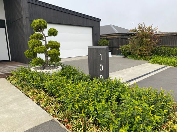 Contemporary residential street frontage with a dark vertical-clad garage facade, broad white garage door, concrete driveway and pedestrian path, landscaped garden beds, and a sculptural topiary tree in a circular planter. A dark address pillar stands prominently near the driveway.