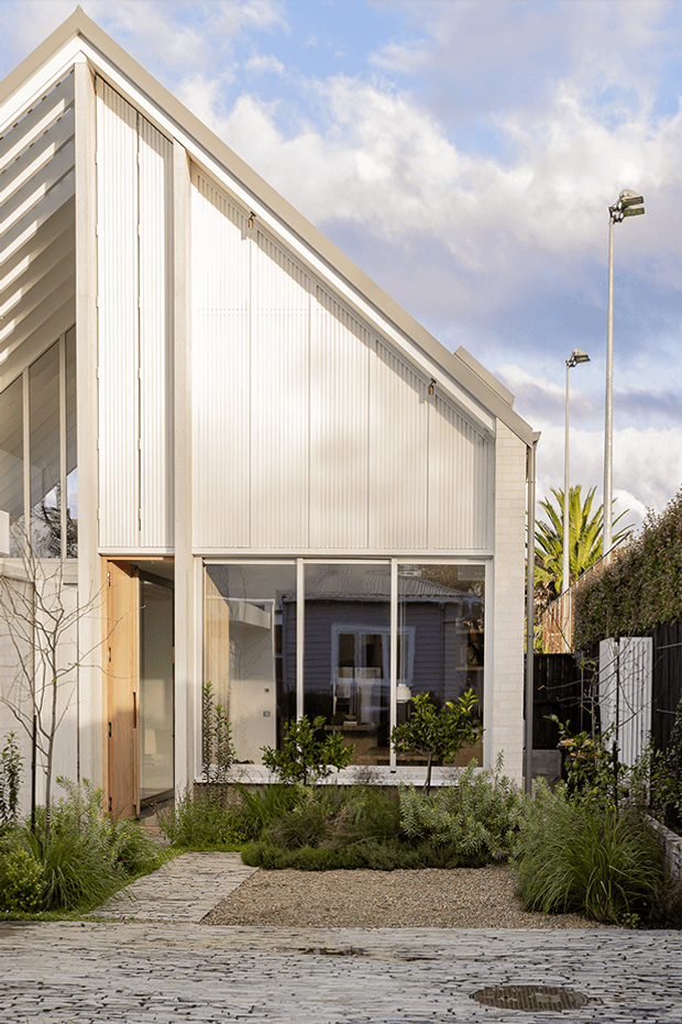 A narrow contemporary residential facade viewed from the street, featuring a steep gabled roof, pale vertical ribbed cladding, exposed light brickwork, tall glazed openings, and a timber entry door. The building is set behind a landscaped gravel and planted frontage with a stone-paved approach. Exterior sports lighting and neighbouring structures are visible in the background under a partly cloudy sky.