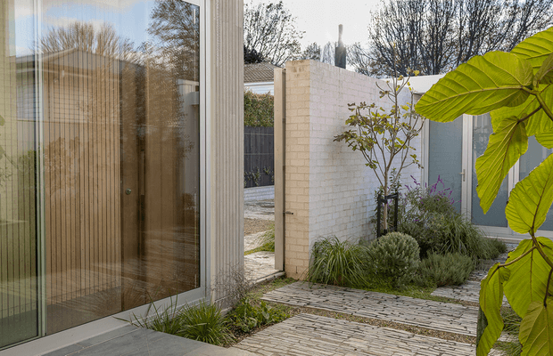 A contemporary courtyard garden viewed from beside a glazed entry or living-space facade. Large glass doors reflect a warm timber-lined interior, while pale brick walls, grey metal fencing, textured stone paving, and layered ornamental planting create a private, refined outdoor threshold.