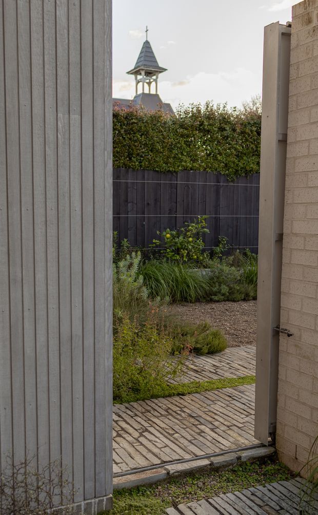 A narrow view through an open gate into a landscaped courtyard or garden. The foreground is framed by a tall vertical timber-clad gate on the left and a pale brick wall with a metal-framed door or gate on the right. Inside are dark timber boundary fencing, dense planting, gravel garden beds, and a patterned brick-paved path. A small cupola-like rooftop structure is visible beyond the hedge.