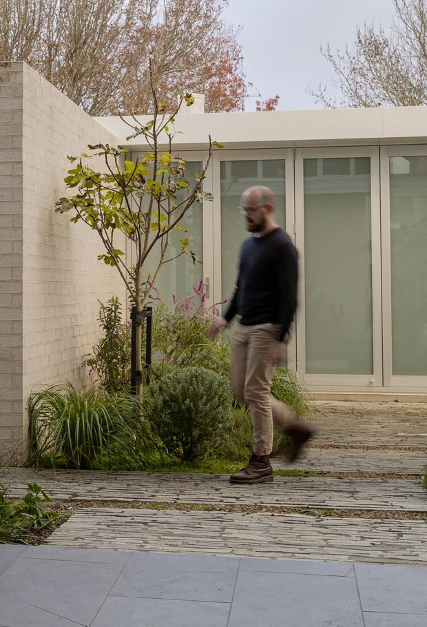 A contemporary landscaped courtyard enclosed by pale masonry walls and a low, flat-roofed glazed building. Full-height frosted glass doors and windows line the facade, opening onto layered stone paving and planted garden beds with a small tree and ornamental grasses. A person is walking through the courtyard.