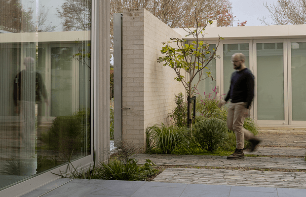 A minimalist contemporary courtyard passage beside a low, flat-roofed building. Large floor-to-ceiling glazed walls with light-coloured frames reflect the planted courtyard, while pale masonry walls, layered stone paving, and landscaped garden beds create a calm connection between the interior and exterior. A person is walking along the path.