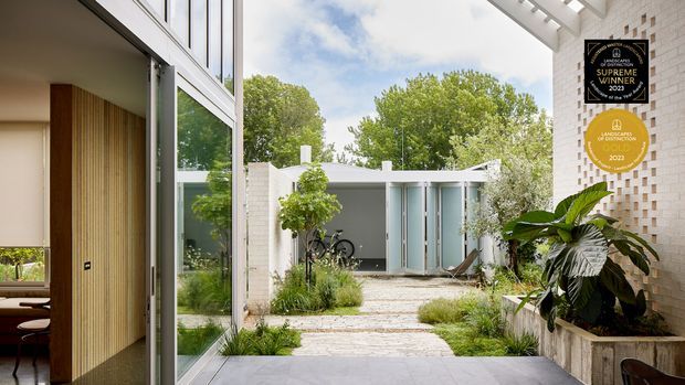 A bright contemporary courtyard viewed from a glazed interior threshold, with pale brick walls, a paved path, landscaped planting, bicycle storage, and a small detached pavilion with glazed doors. Vertical timber screening and expansive glazing frame the left side, while award plaques are mounted on the right wall.