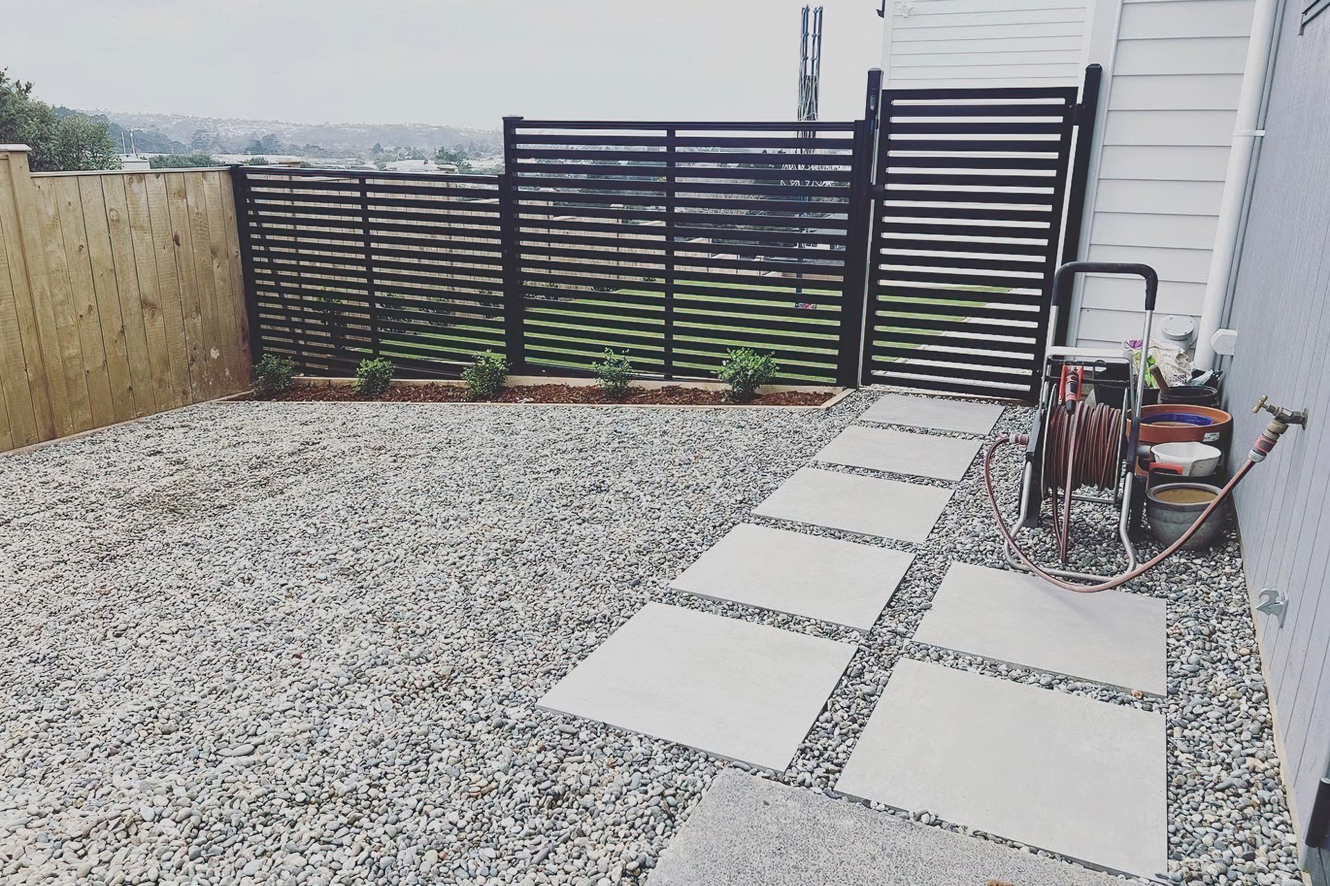 Modern black fence and gate that divide the yard from a pebble area with white stone tiles to walk on
