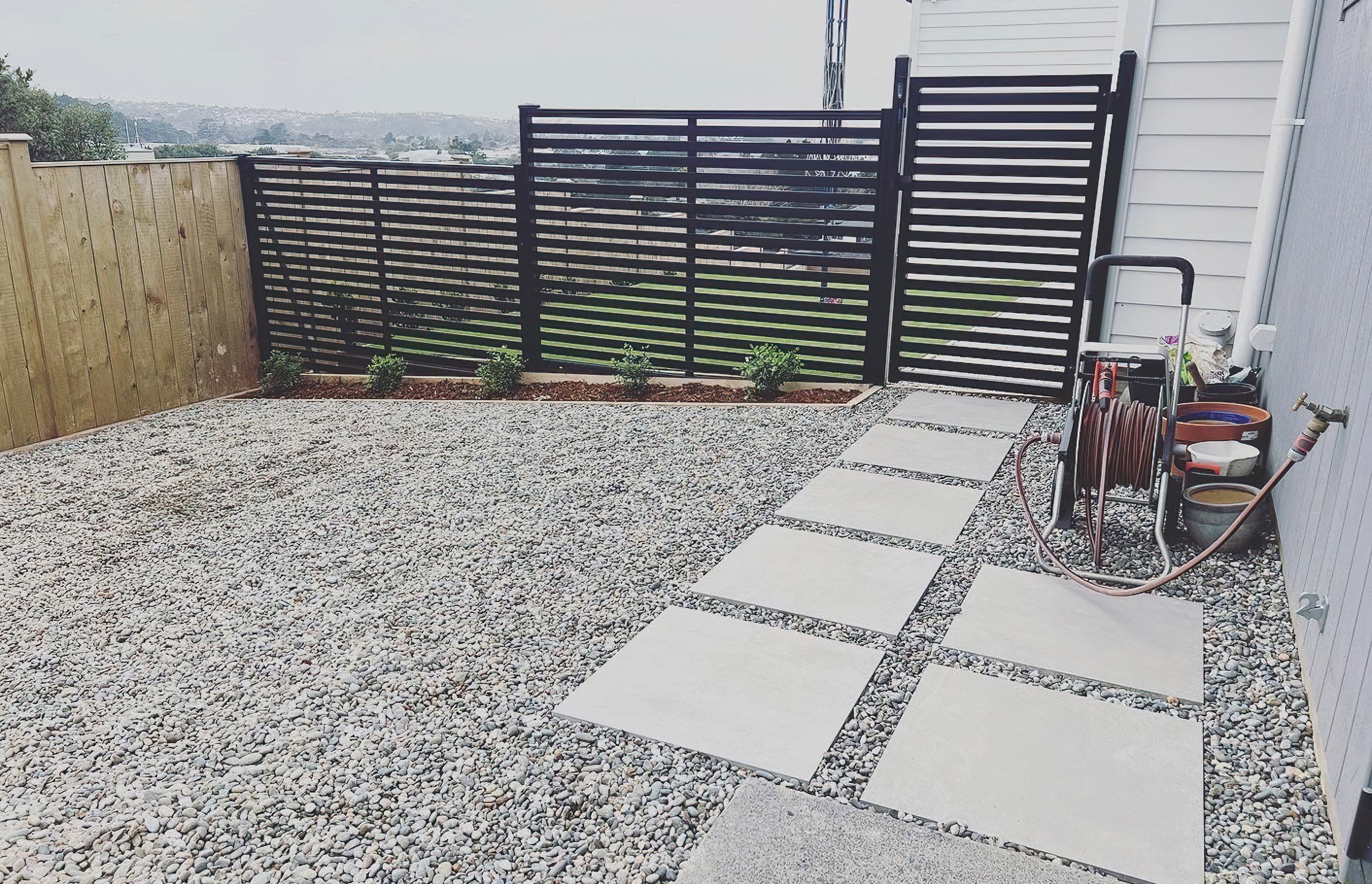 Modern black fence and gate that divide the yard from a pebble area with white stone tiles to walk on