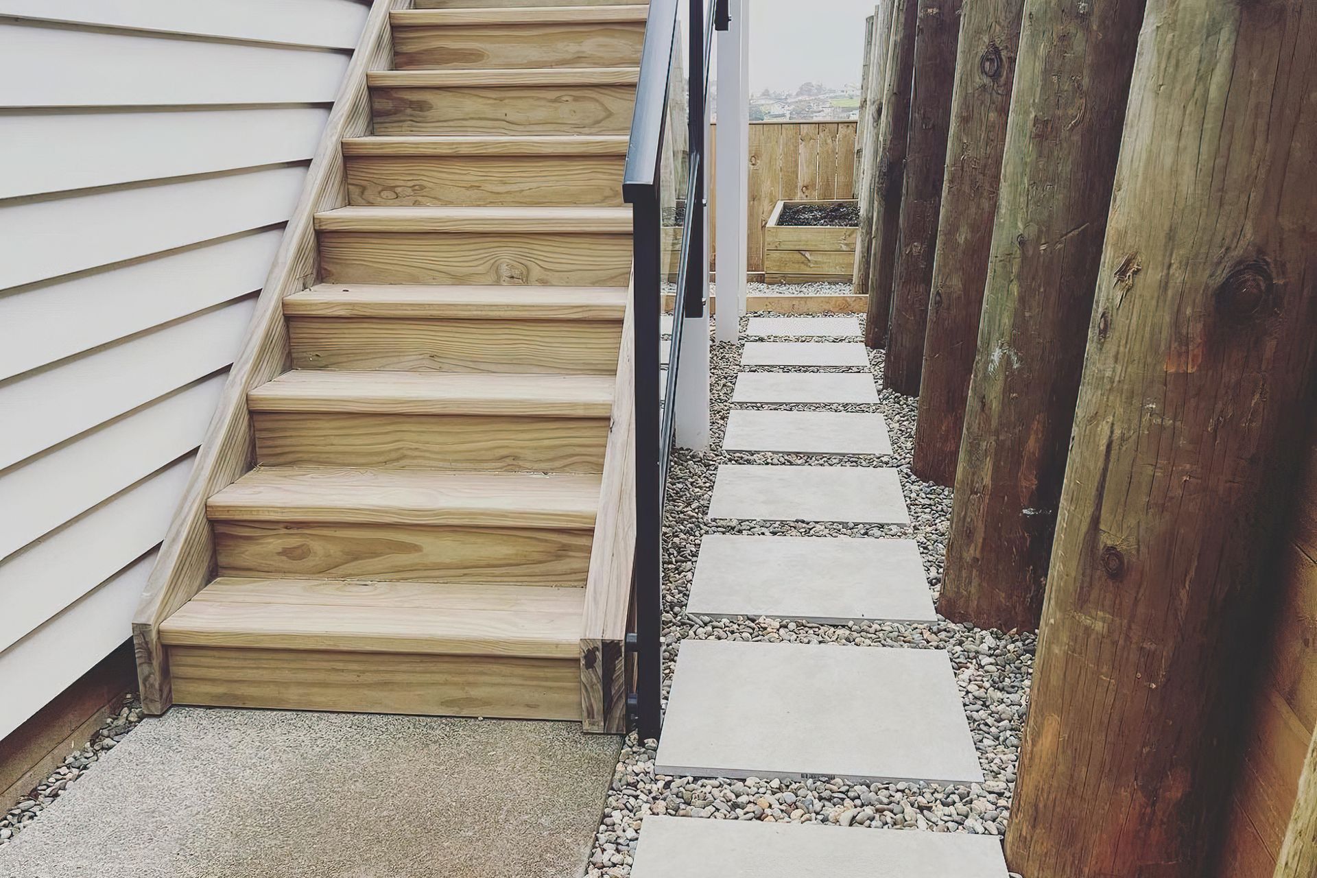 Wooden stairway next to a path to the backyard made with concrete tiles surrounded by pebbles