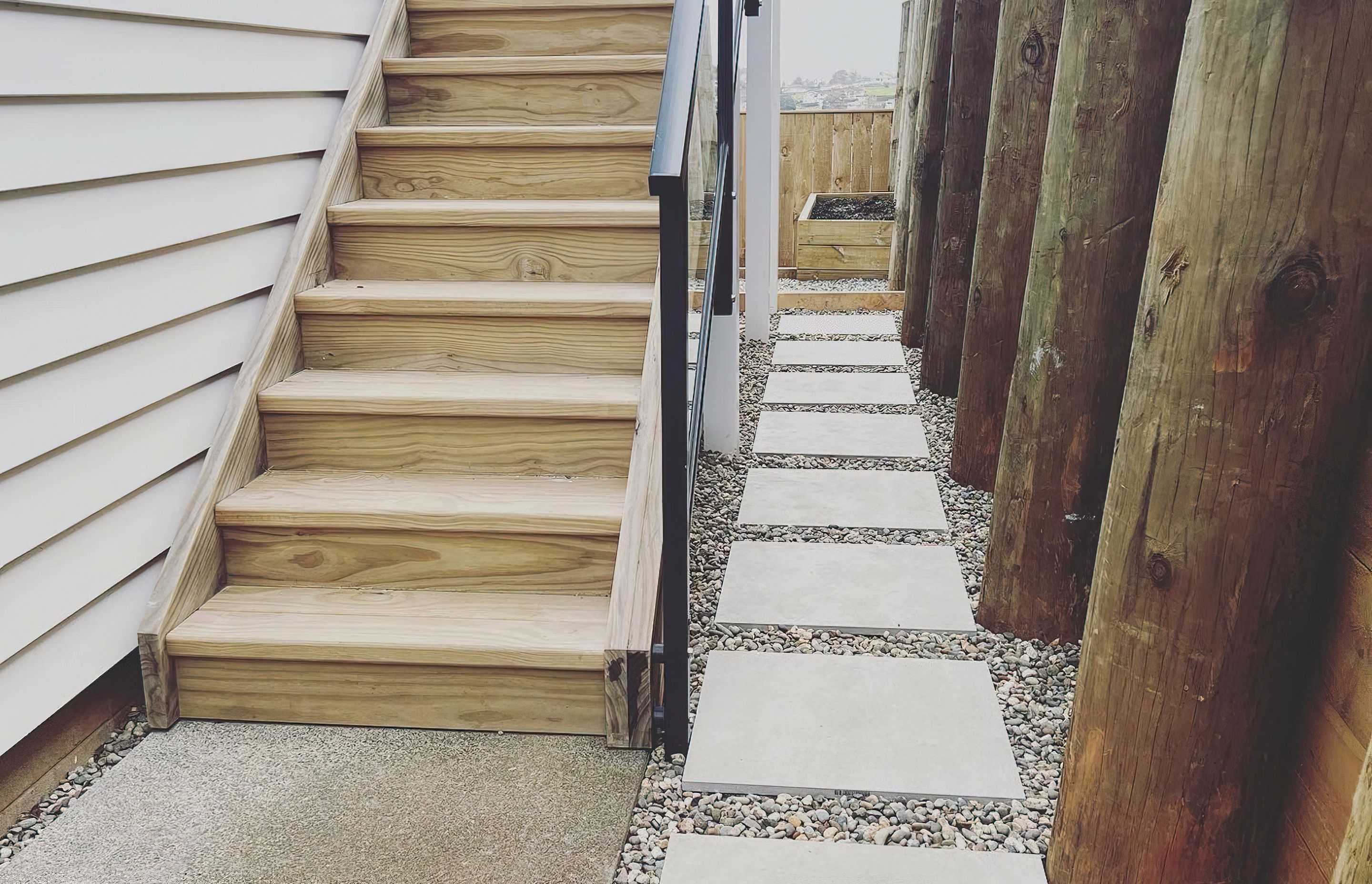 Wooden stairway next to a path to the backyard made with concrete tiles surrounded by pebbles