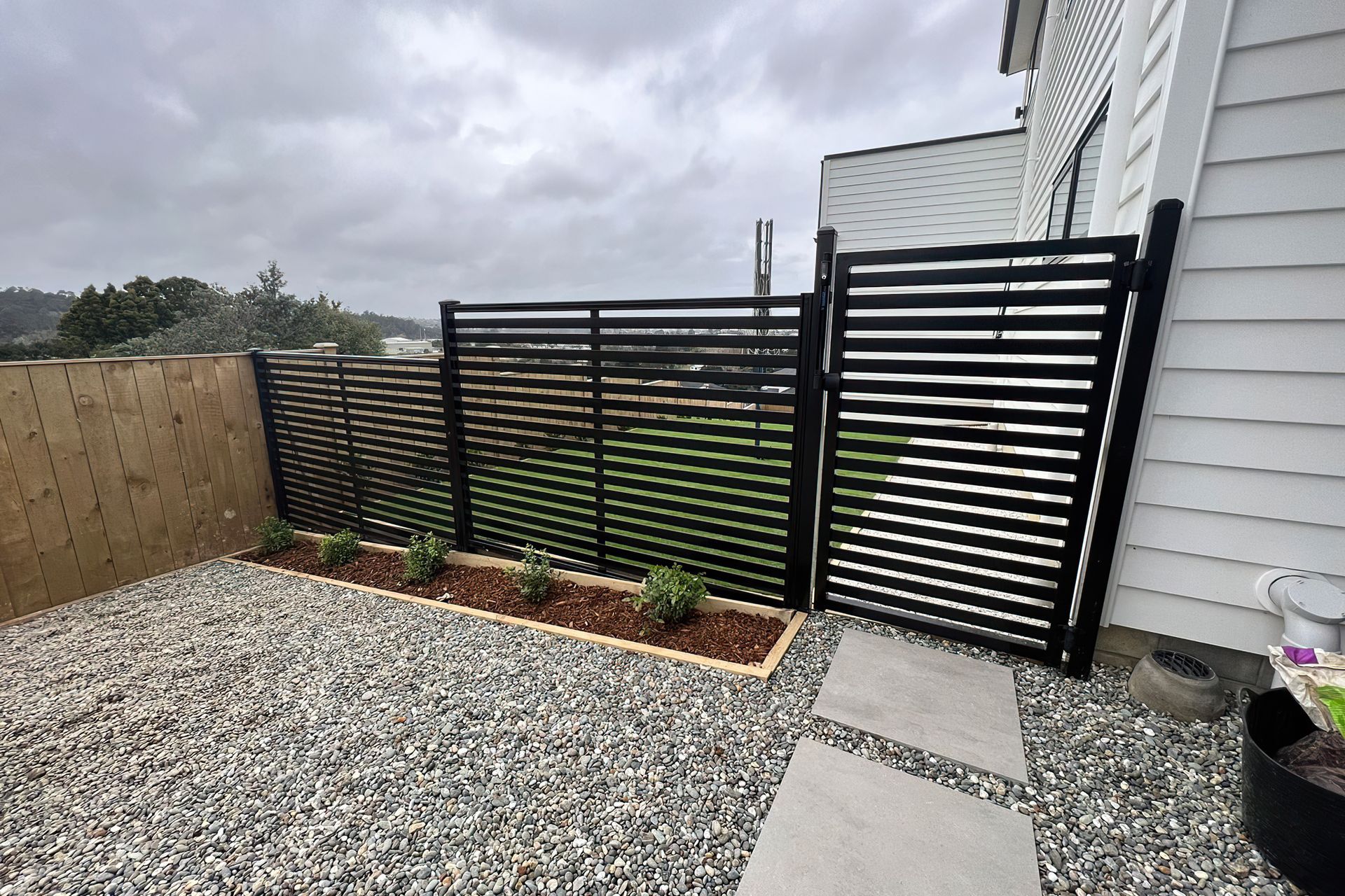 Modern black fence and gate that divide the yard from a pebble area with white stone tiles to walk on