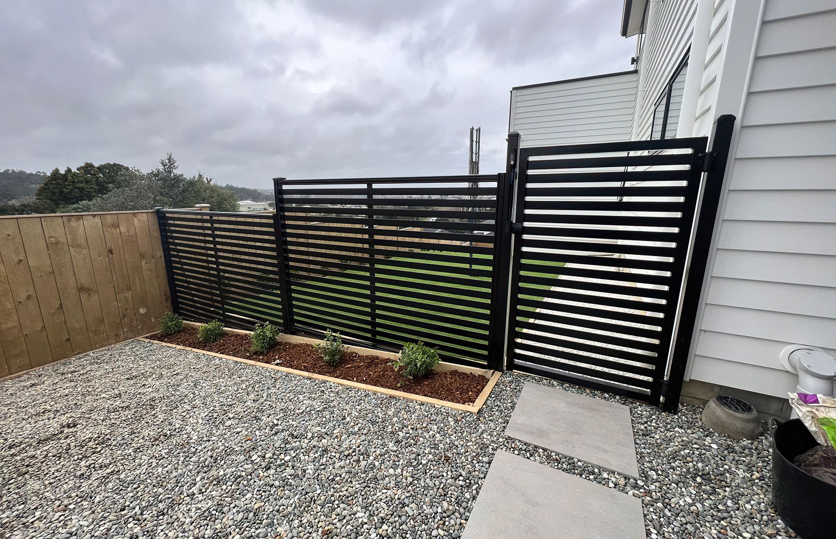 Modern black fence and gate that divide the yard from a pebble area with white stone tiles to walk on