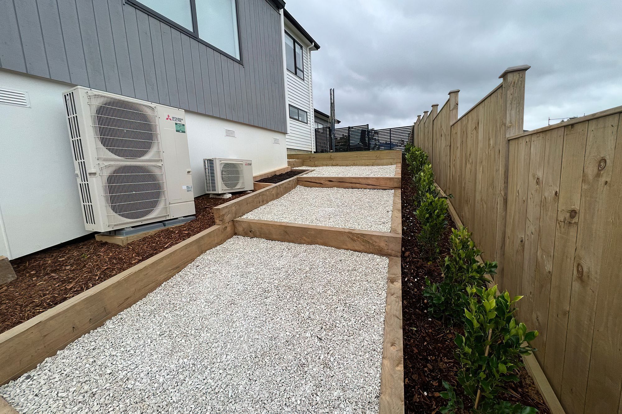 Three large steps going down the side of a newly built home and a row of freshly planted trees along the fence line