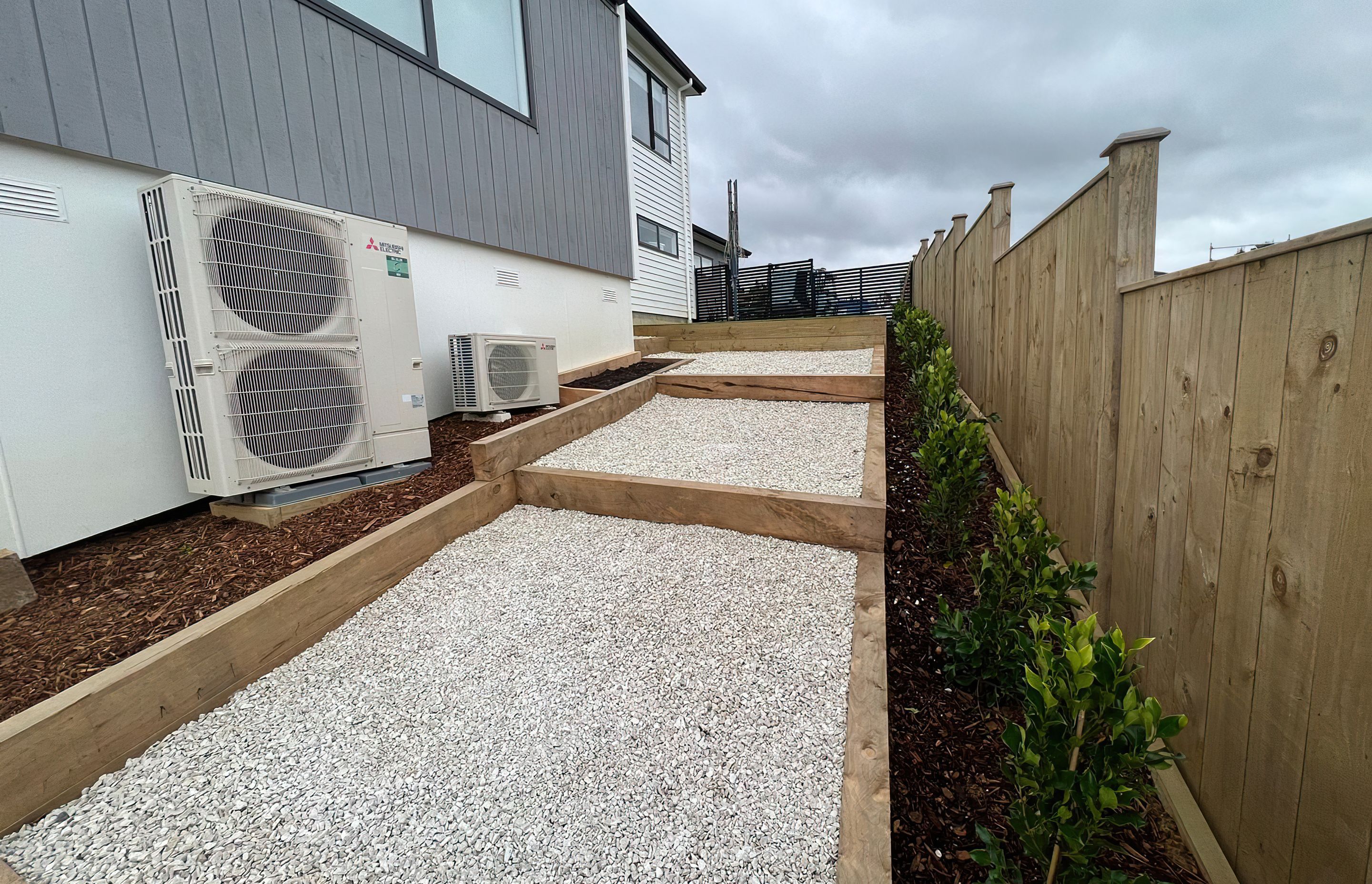 Three large steps going down the side of a newly built home and a row of freshly planted trees along the fence line