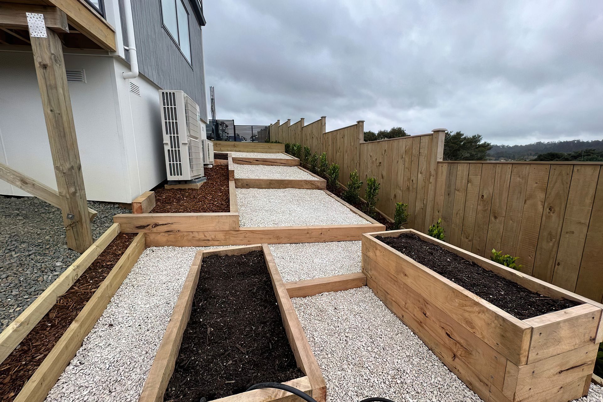 Large outside area going down the side of a newly built home with a couple vegetable beds and a row of freshly planted trees along the fence line