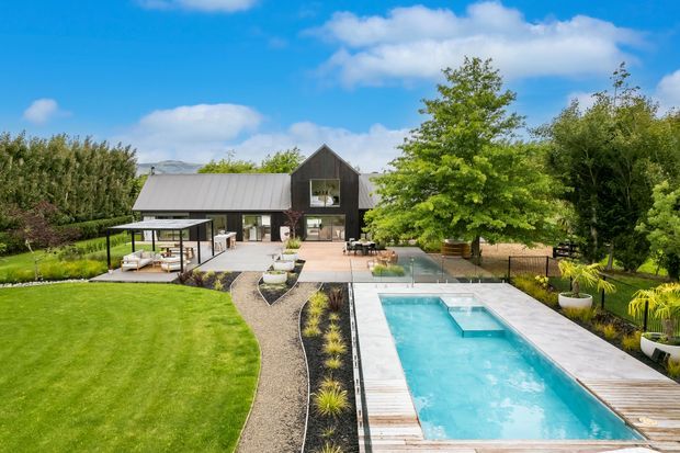 A landscaped residential property viewed from the garden, featuring a dark-clad gable-roofed house, expansive lawn, outdoor entertaining area, paved terrace, and a long rectangular swimming pool with timber decking. Mature trees and structured planting surround the contemporary rural-style setting, with hills visible in the distance.