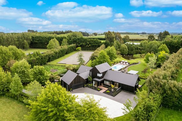 Aerial view of a large contemporary rural property surrounded by mature trees, manicured lawns, hedges and open farmland. The dark-clad residence has multiple pitched roof volumes, broad paved driveways and terraces, with a visible swimming pool and outdoor seating area behind the house.